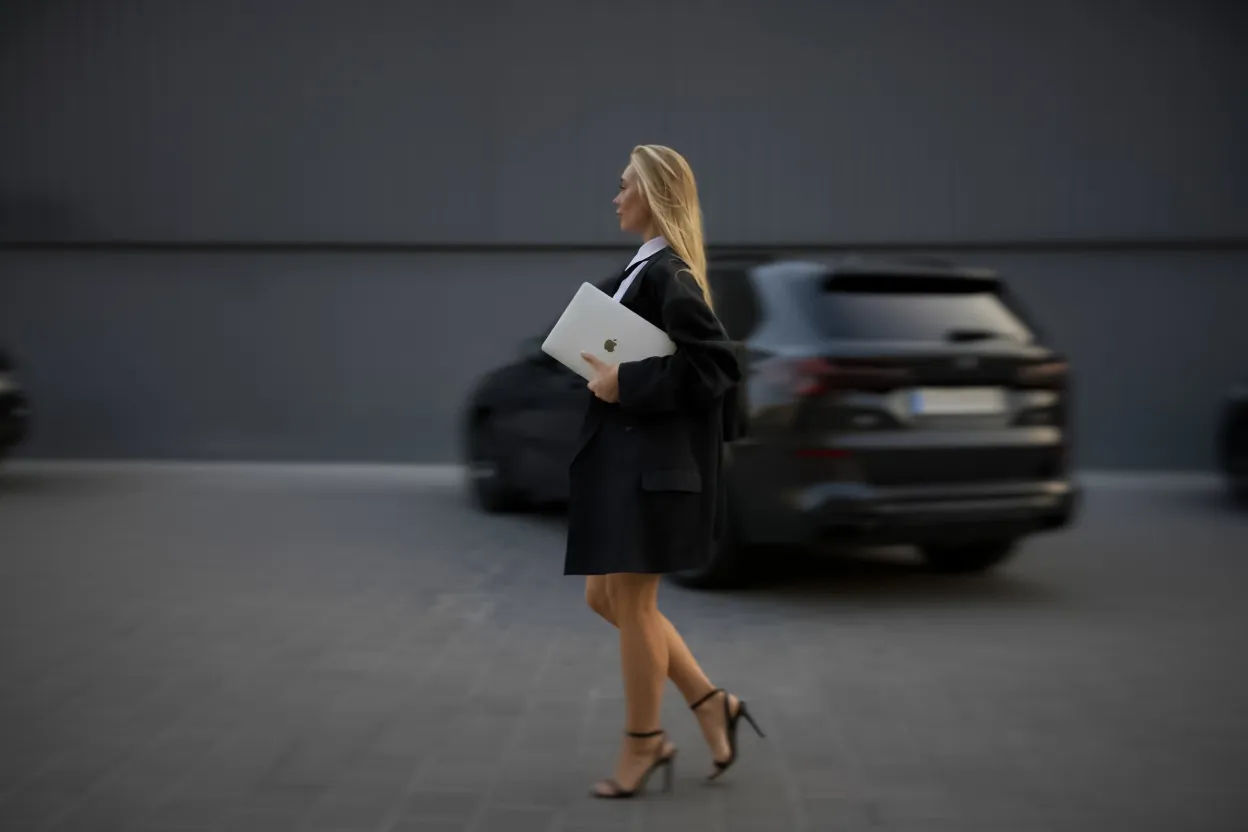 Kamilla walking past a black SUV with a laptop